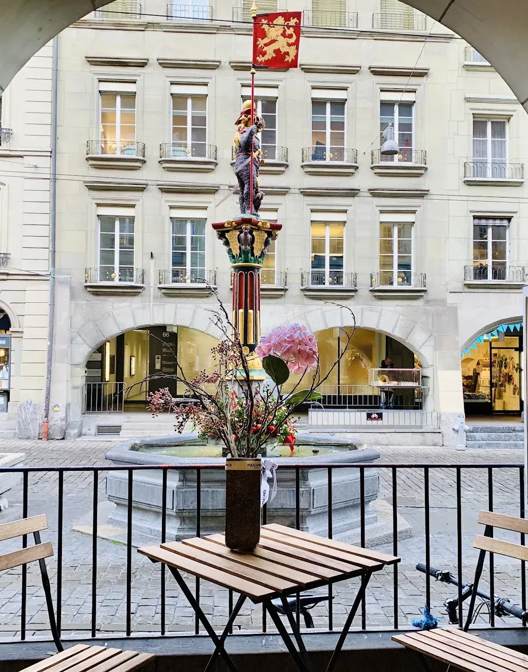 A café table under a stone arcade in Bern, looking out at a painted fountain statue