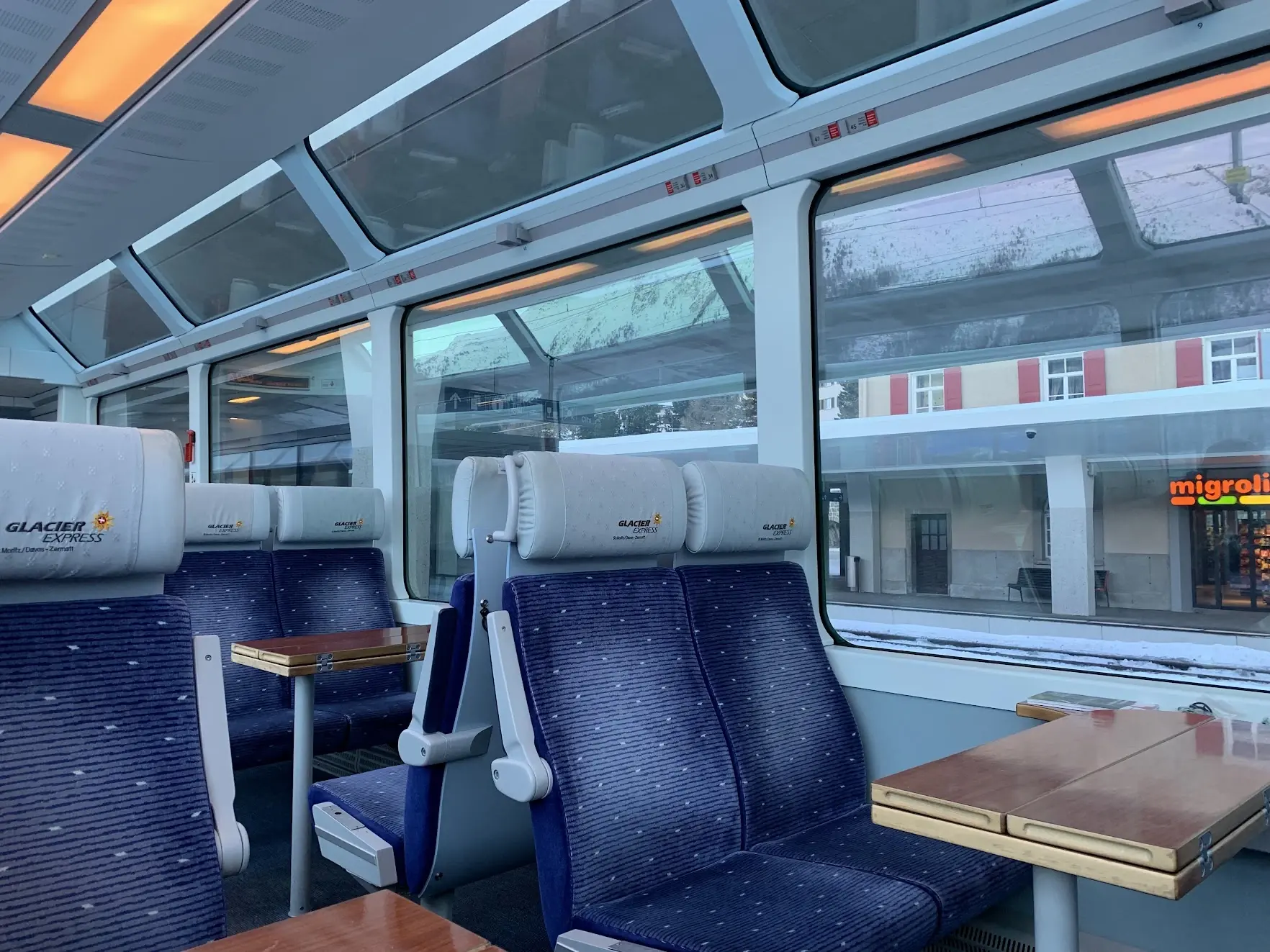 Inside a Glacier Express panoramic carriage: blue seats, small tables and windows that curve up into the roof
