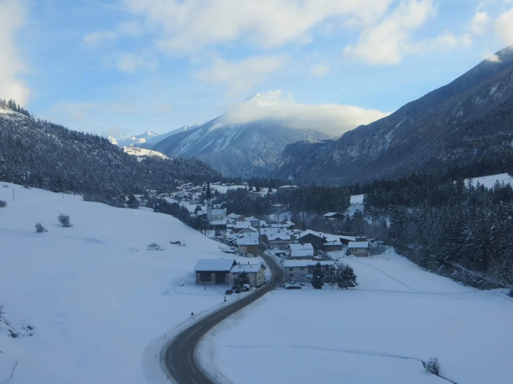 A winding road leading to a snowy village with a church spire, below cloud-capped mountains