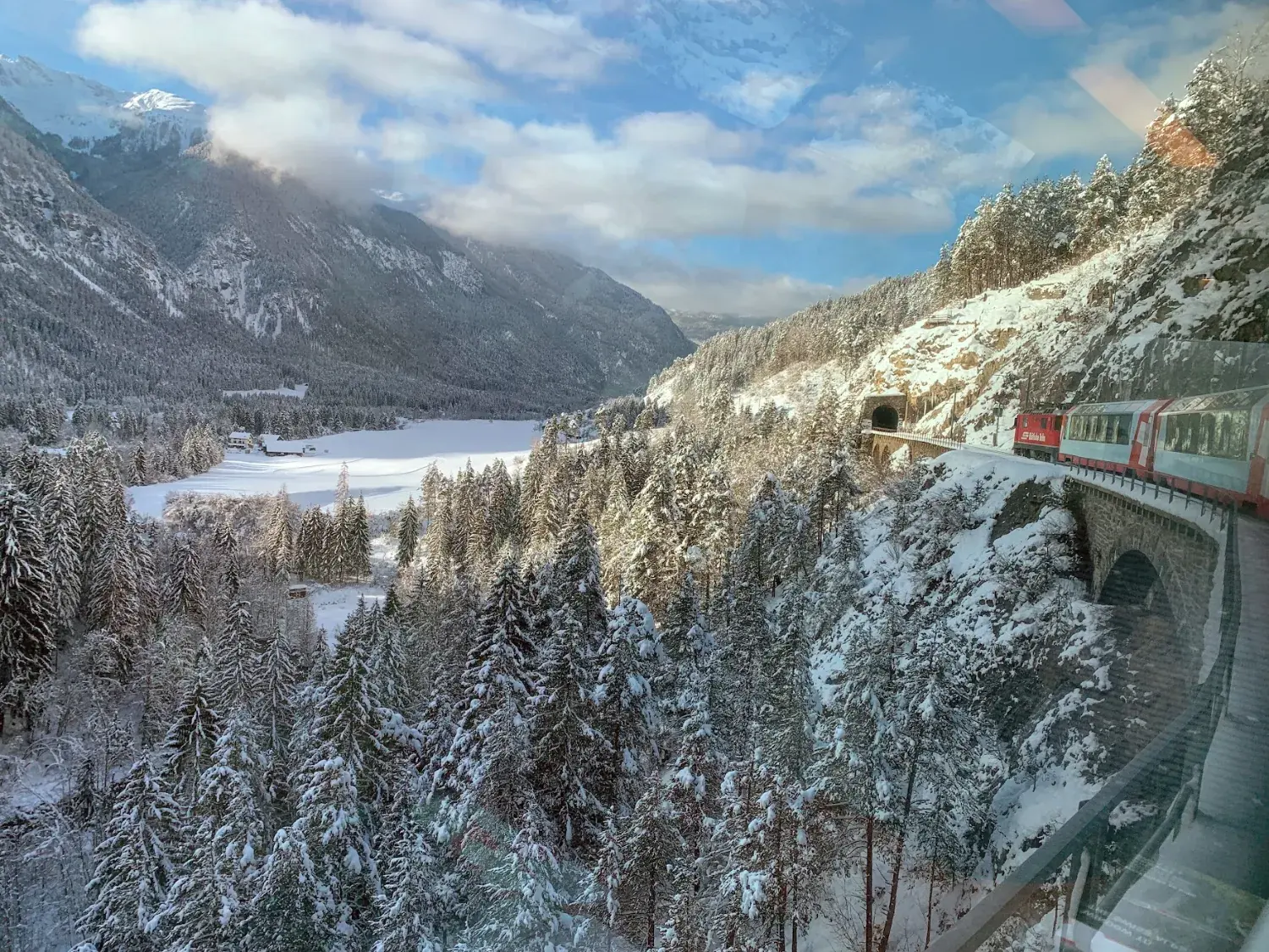 The red Glacier Express crossing a stone viaduct above a snowy forest and valley, seen from a window of the same train