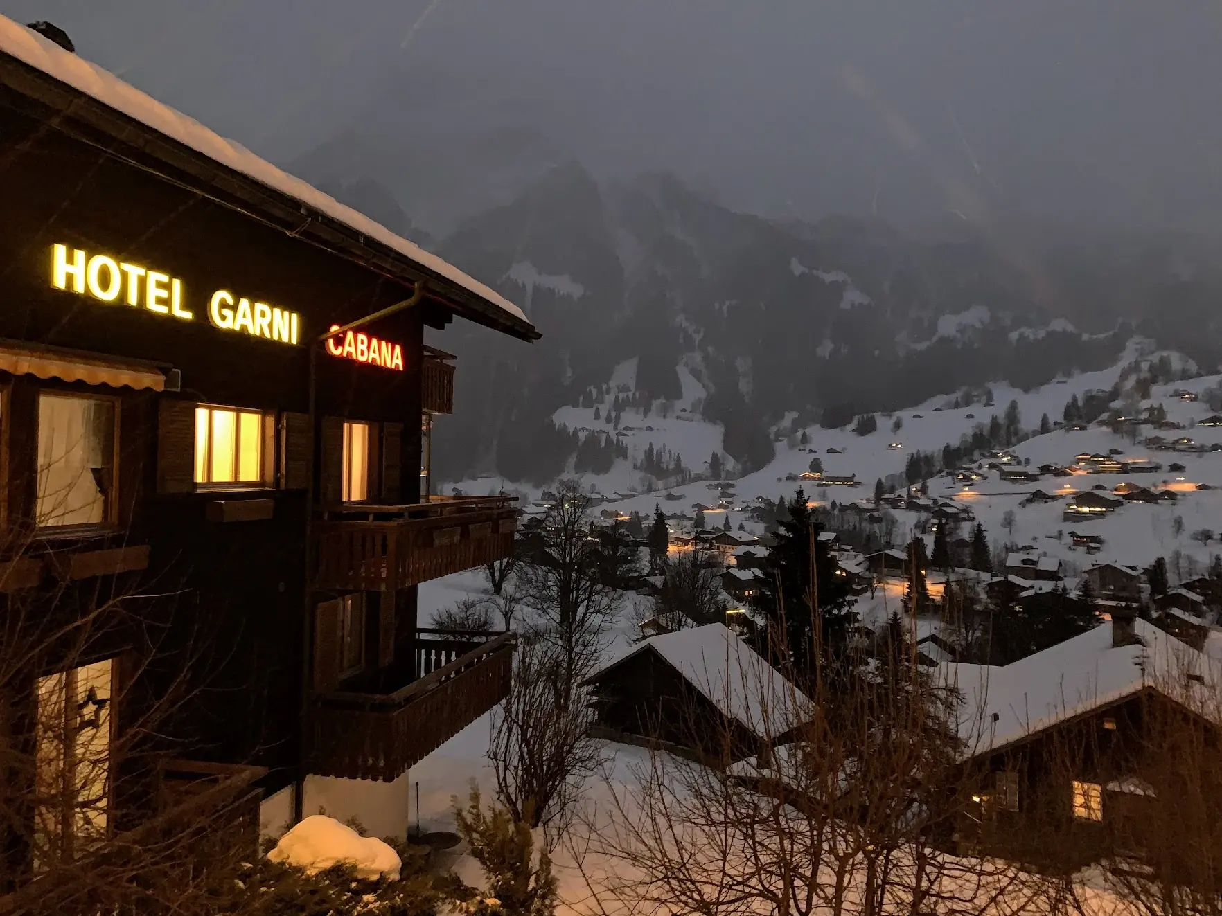 The lit sign of Hotel Garni Cabana on a dark wooden chalet at dusk, above the snowy lights of Grindelwald