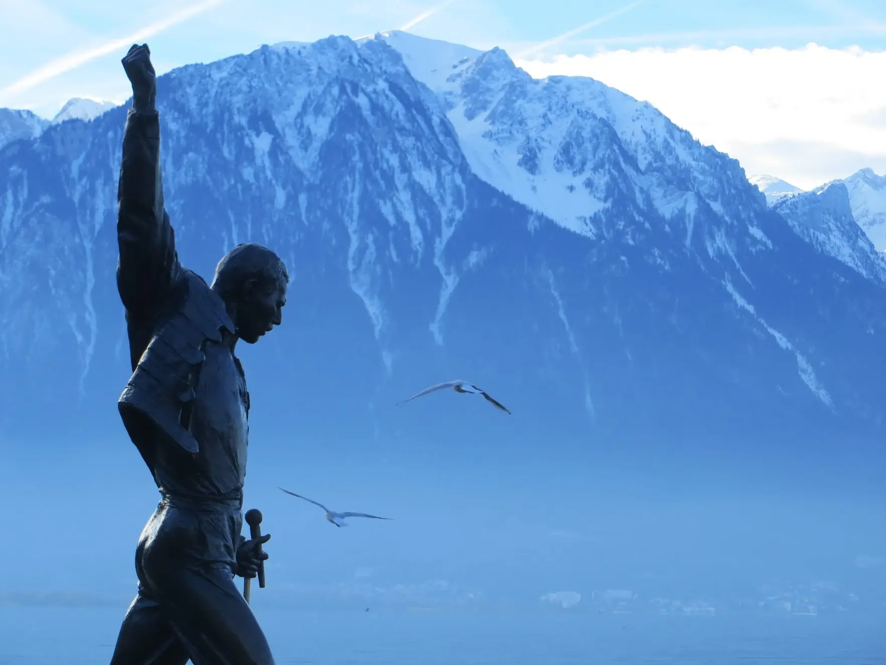 The statue of Freddie Mercury with his fist raised, in silhouette against Lake Geneva and snowy mountains, with seagulls