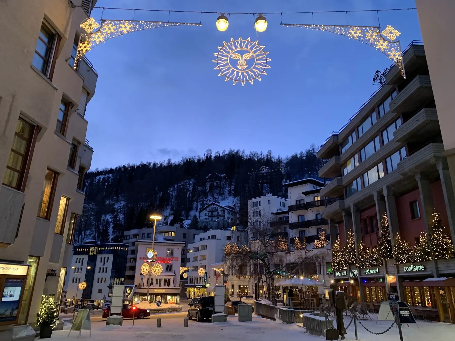 St. Moritz at dusk with a glowing sun-shaped Christmas light over the street and the Hauser hotel and café lit up