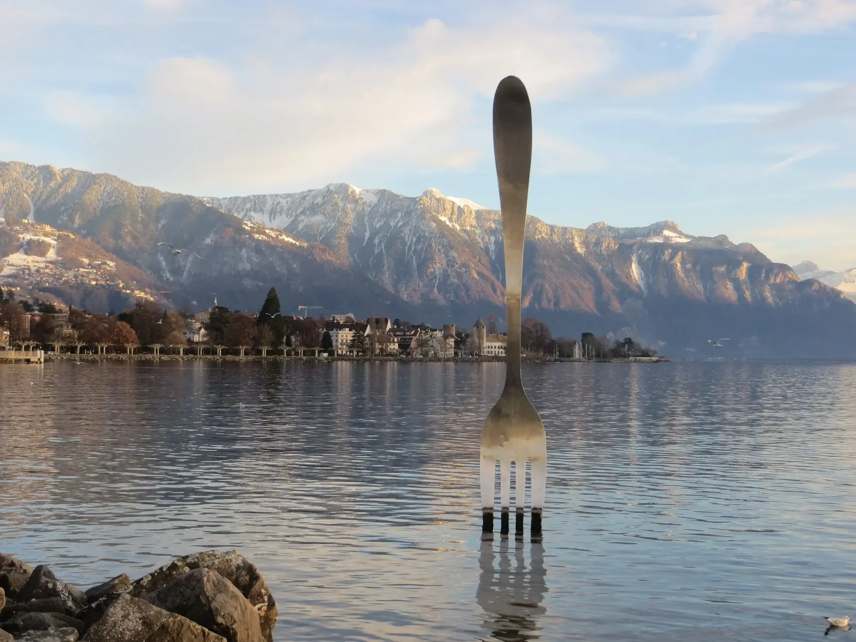 A giant stainless-steel fork standing in Lake Geneva at Vevey, with the town and mountains behind
