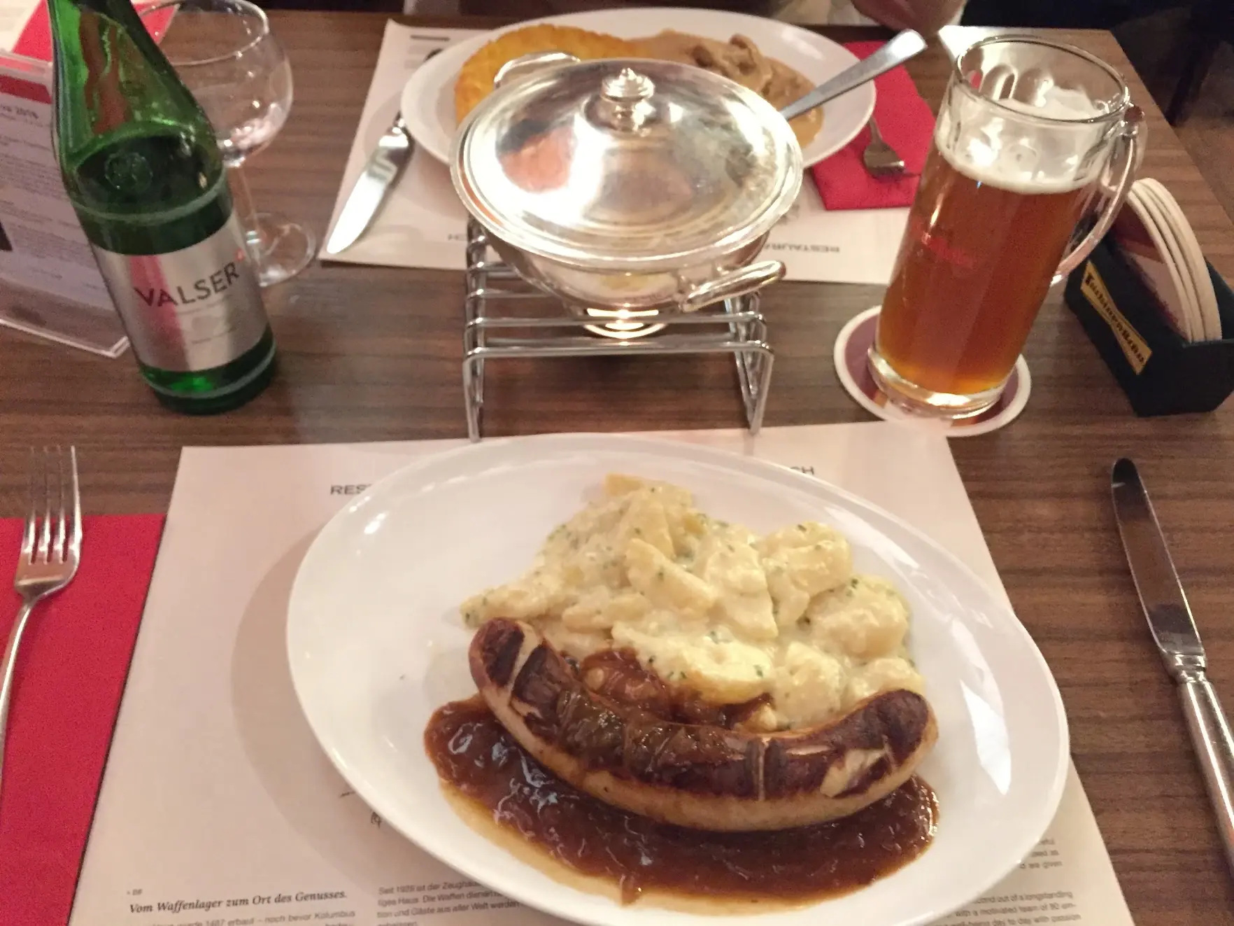 A bratwurst with onion sauce and creamy potatoes, with rösti and a beer behind