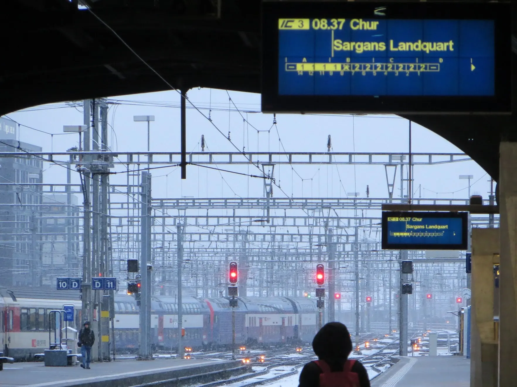 A departure board at Zurich station for the 8:37 train to Chur, with snow falling over the tracks