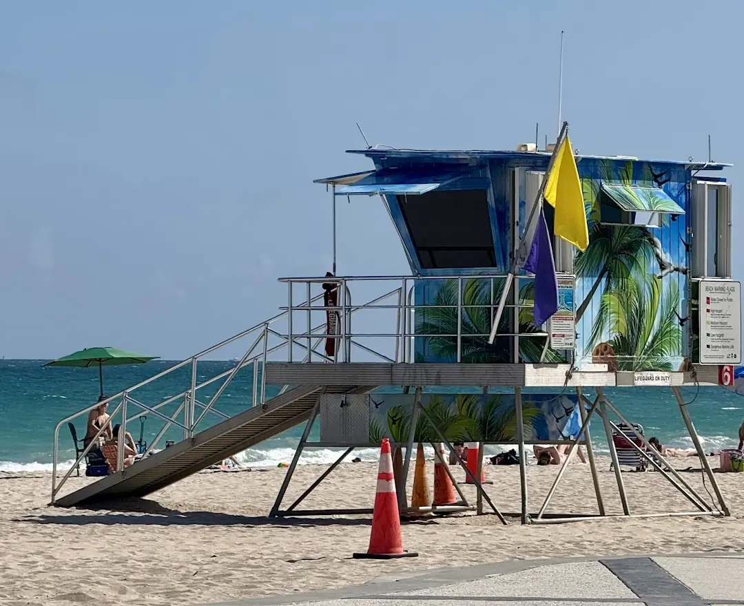 A blue lifeguard tower painted with palm trees on Fort Lauderdale beach, with a yellow flag flying