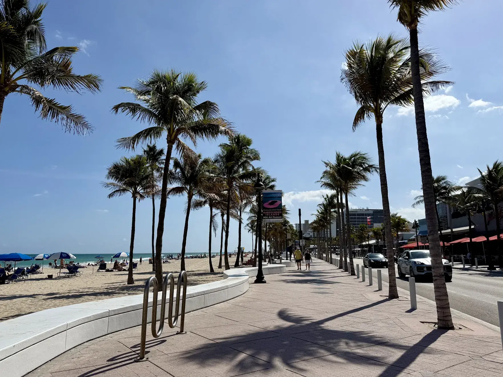 A palm-lined beachfront promenade along Fort Lauderdale beach, with the ocean on the left and the road on the right