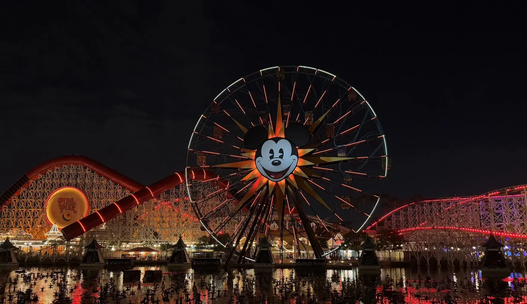 The Mickey Ferris wheel and Incredicoaster lit up at night, reflected in the lagoon