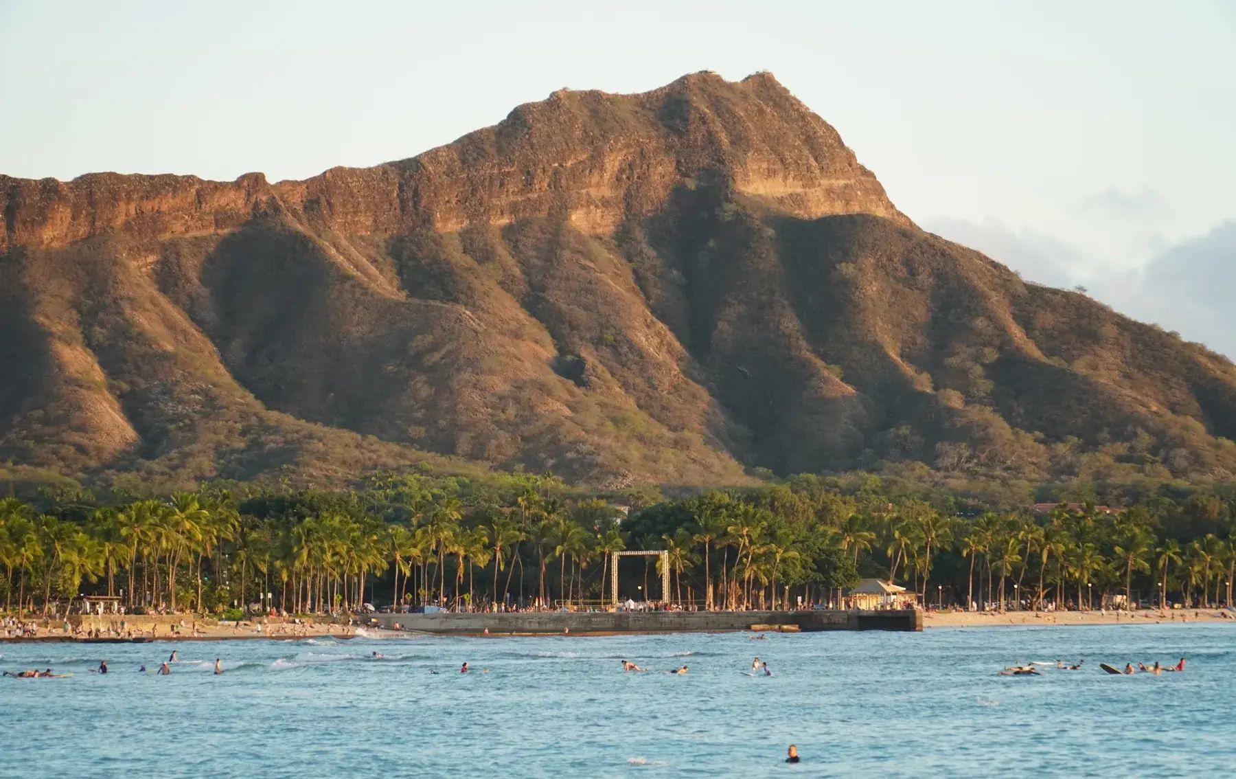 Diamond Head lit by late-afternoon sun, behind a line of palm trees and surfers in the water