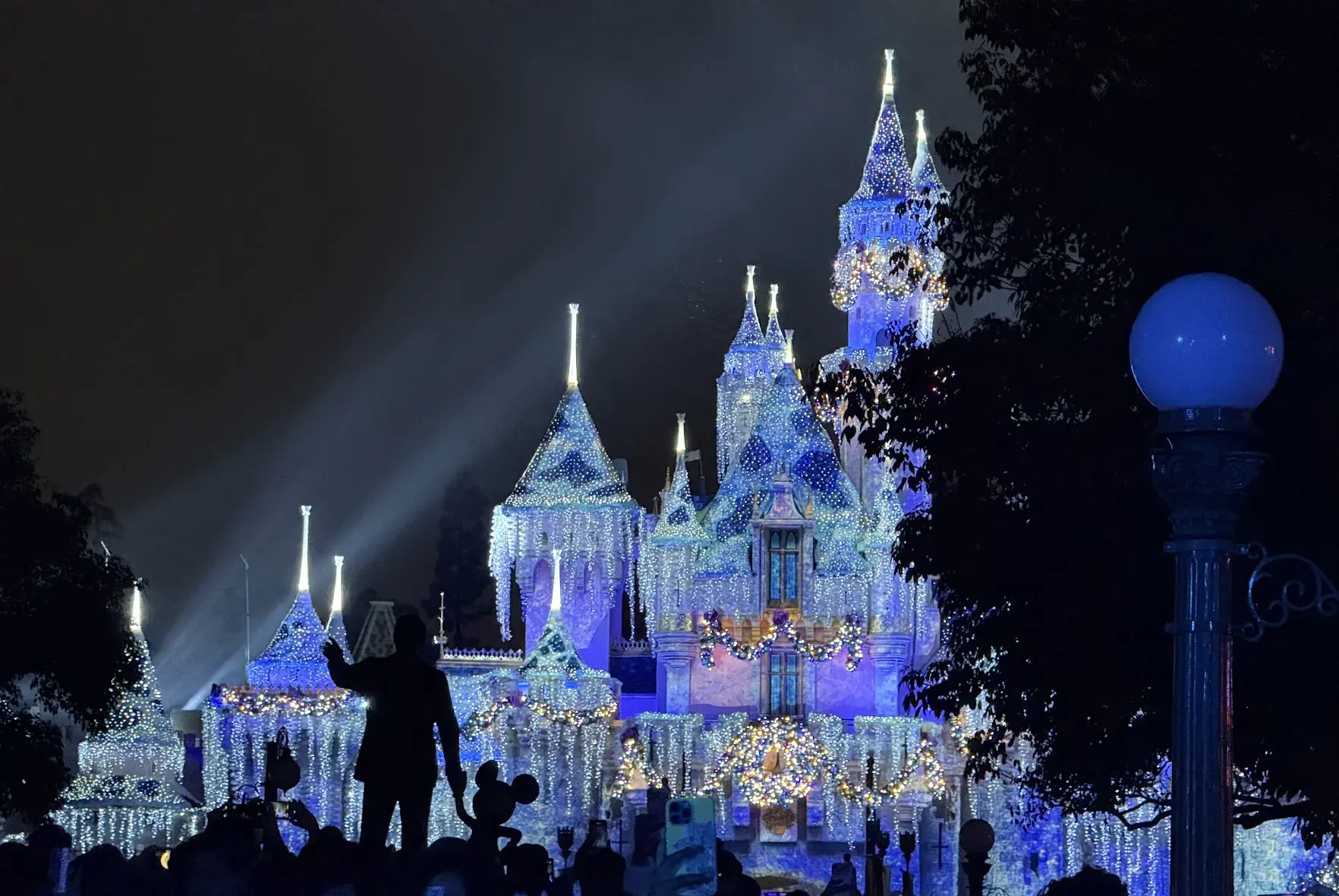 Sleeping Beauty Castle covered in icicle lights at night, with the Partners statue in silhouette