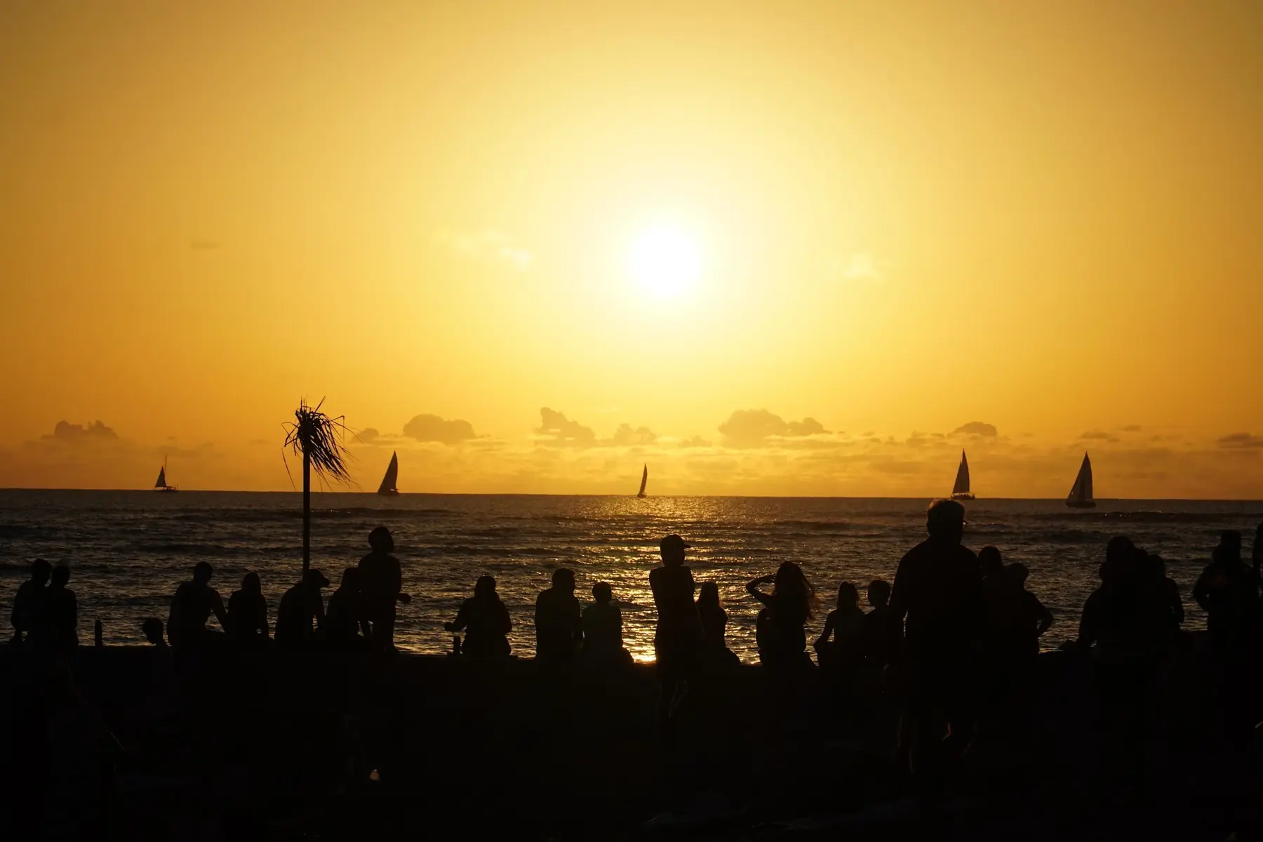 Silhouettes of people on Waikiki Beach watching the sun set over the ocean, with sailboats on the horizon